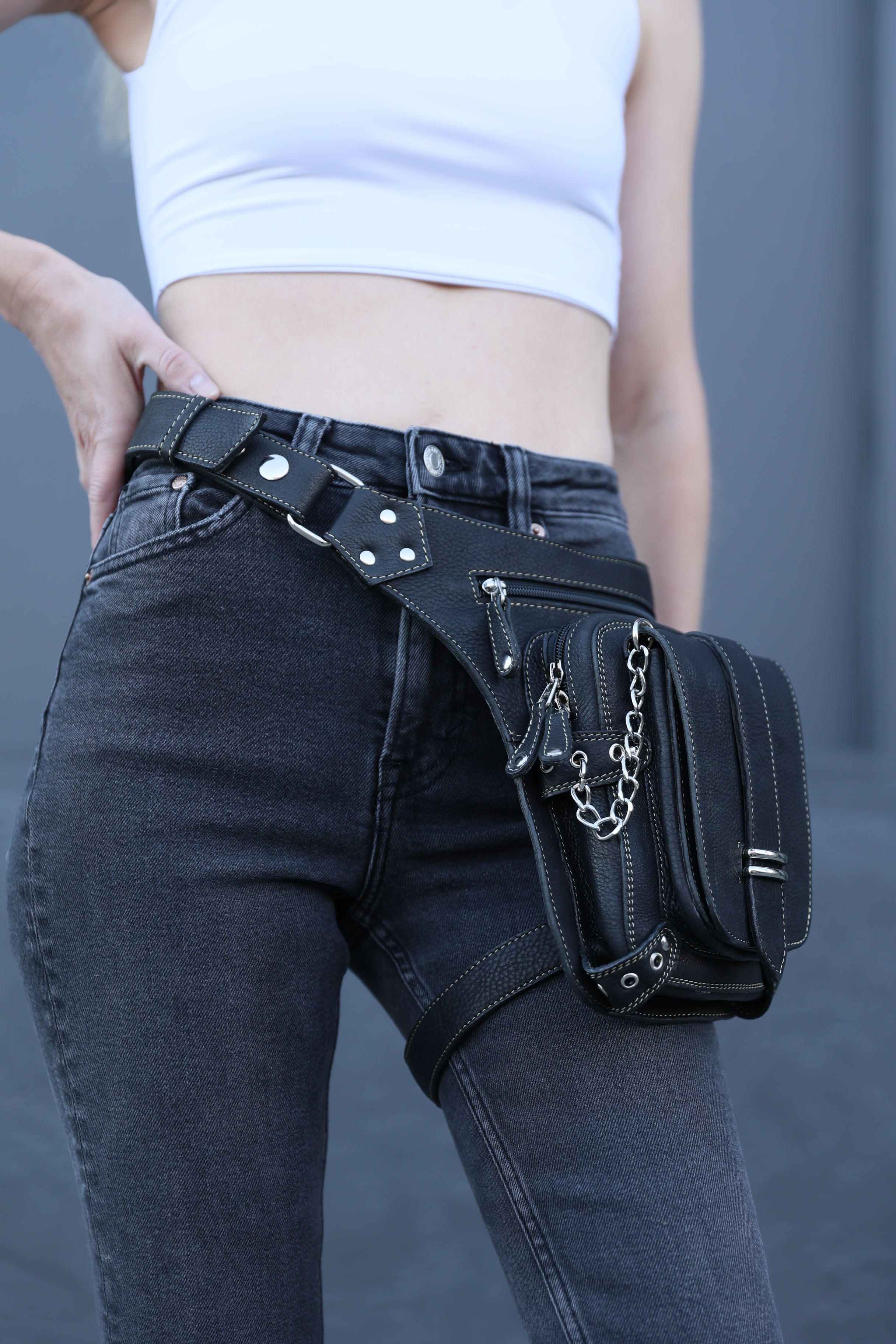 Person wearing a black leather waist bag with studs and chains on a gray background