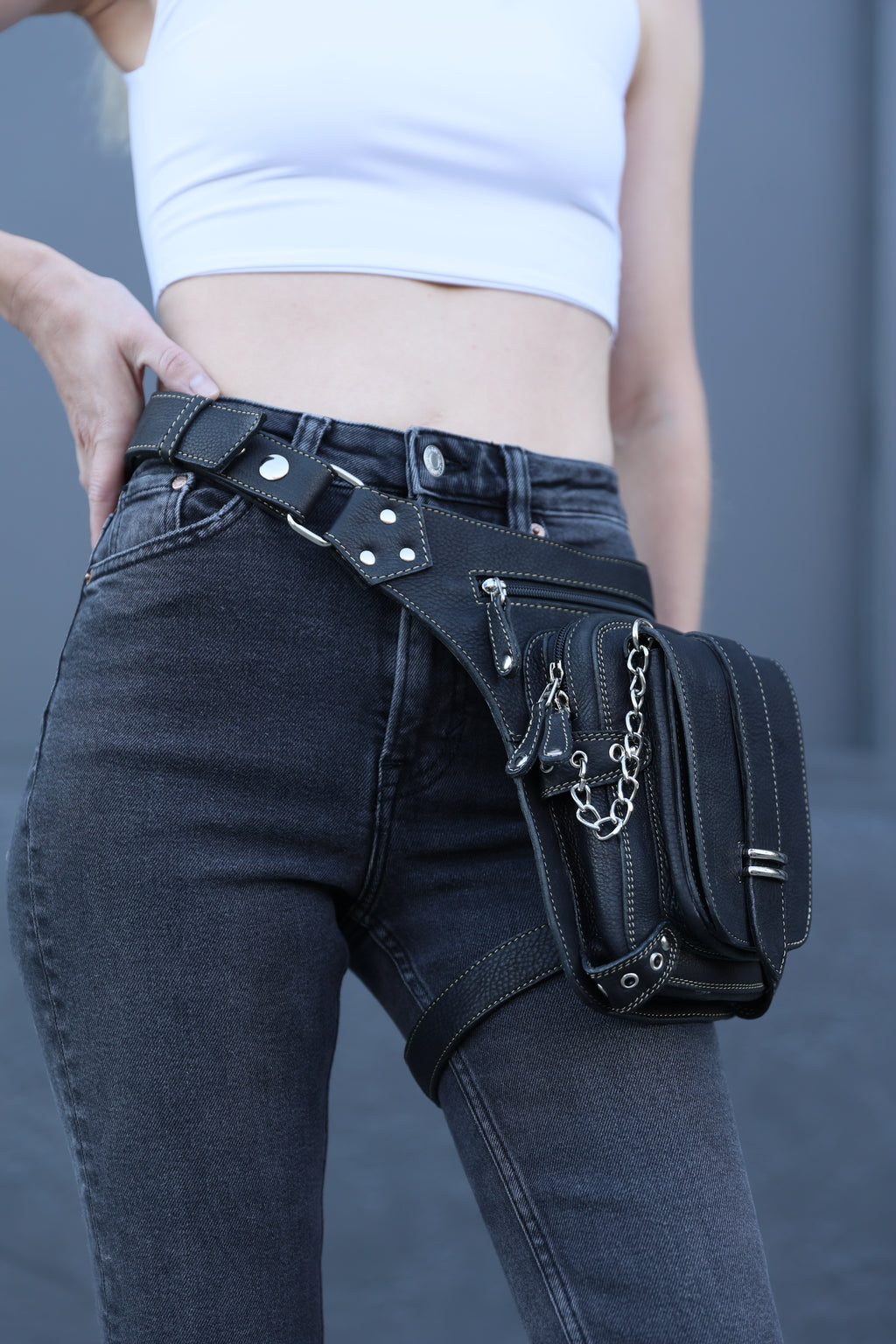 Person wearing a black leather waist bag with studs and chains on a gray background