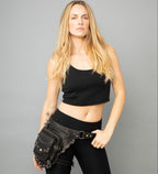 Woman wearing a black crop top and black pants with a studded leather  leg bag on a gray background