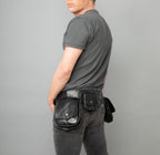 A man facing away from the camera is wearing a black leather utility belt with large side pouches and a small back pouch. He has a grey shirt and blue jeans on.