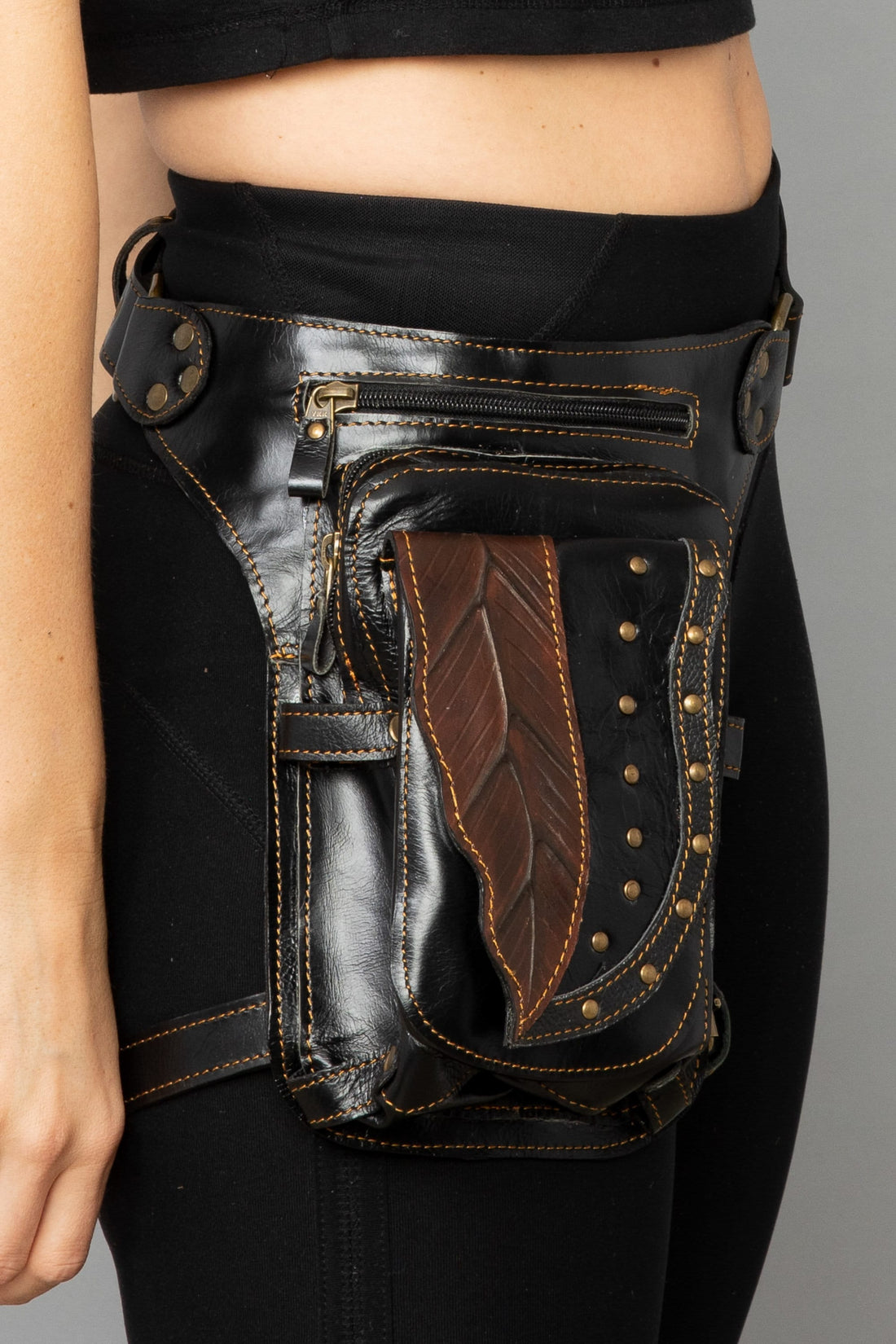 A person wearing a black leather belt bag with a decorative leaf detail and a brown leaf design, featuring multiple zippered compartments and a removable leg strap.